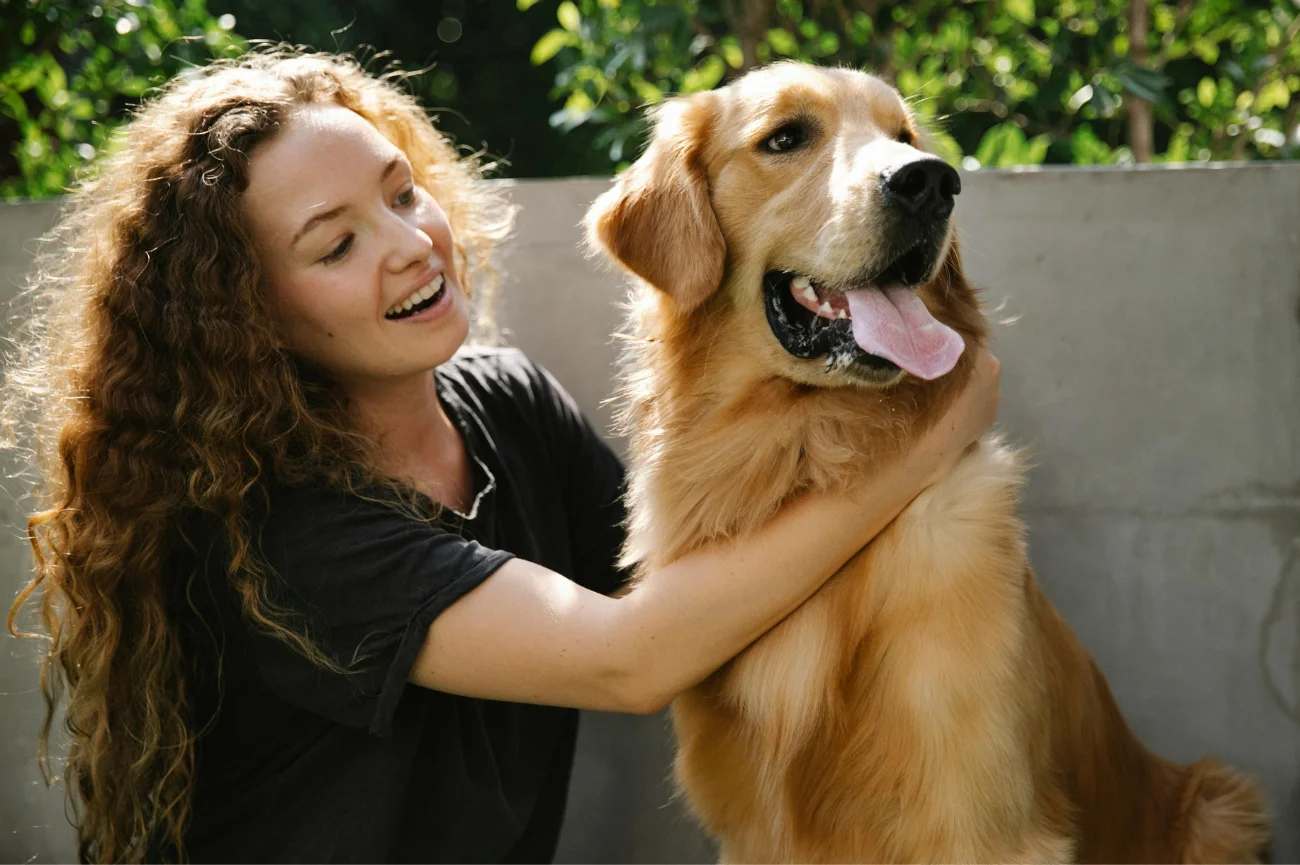 Młoda kobieta z długimi, brązowymi, kręconymi włosami uśmiecha się i obejmuje golden retrievera