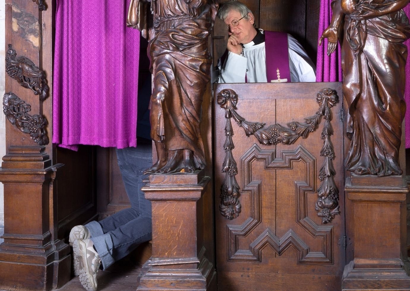 Mężczyzna klęczy w drewnianym konfesjonale, jest zasłonięty różową kotarą, widać jedynie nogi. W konfesjonale siedzi też ksiądz w okularach, który słucha spowiednika